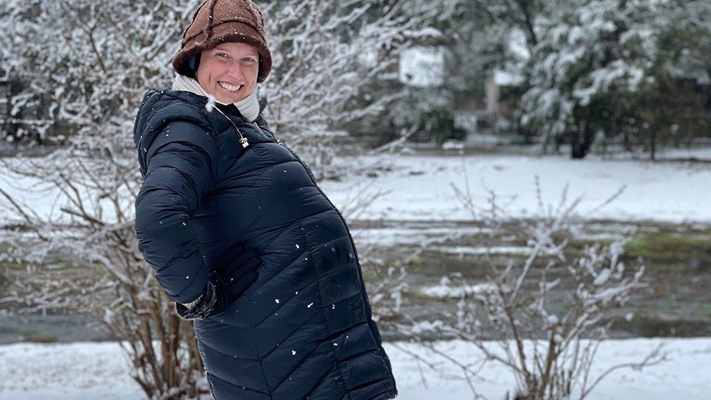 穿着她的冬天外套和帽子的三重阴性炎症乳腺癌幸存者alessandra陆地在雪中，当展示她怀孕的腹部时